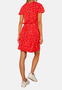 Robe rouge à petit imprimé floral blanc, manches courtes et taille ceinturée. Portée avec des baskets blanches. Tissu lisse et coupe décontractée.