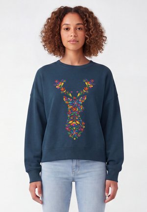 Jeune femme aux cheveux bouclés portant un sweat-shirt bleu marine avec un motif coloré de tête de cerf floral et un jean bleu clair sur un fond uni.