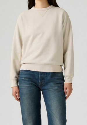 Sweat-shirt beige à manches longues avec poignets et bas côtelés porté avec un jean bleu sur une personne debout.