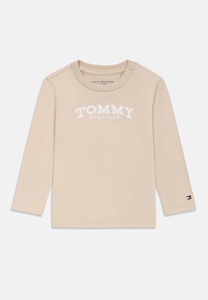 Beżowa koszulka z długim rękawem dla malucha z napisem „Tommy Hilfiger” z przodu i naszywką z logo na mankiecie lewego rękawa, z zatrzaskami na ramieniu.