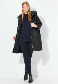 Černý prošívaný kabát s kapucí a zlatými kovovými detailami, vrstvený přes tmavě modrý top a tailored kalhoty, doplněný černými kotníkovými botami.