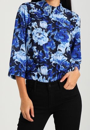 Kwiecista bluzka z wysokim kołnierzem i rękawami do połowy łokcia. Charakteryzuje się wzorem niebieskich i białych kwiatów na ciemnym tle. Noszona z czarnymi spodniami.