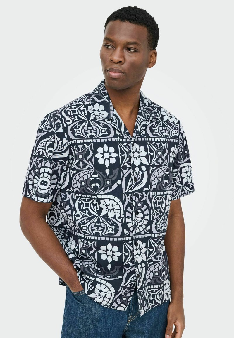 Hombre con camisa de botones de manga corta negra y blanca con estampado floral y jeans azules, mirando hacia un lado con una mano en el bolsillo.