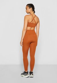 Reggiseno sportivo arancione con spalline incrociate abbinato a leggings arancioni a vita alta. Il modello indossa scarpe da ginnastica nere con dettagli bianchi.