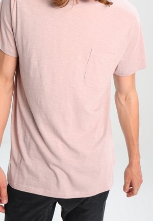 Mand iført en lyserød kortærmet t-shirt med en lille brystlomme og mørke bukser, stående mod en ensfarvet baggrund.