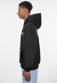 Sweatshirt à capuche noire fabriqué dans un tissu doux, avec une poche avant et des poignets côtelés. Le design est simple et sans ornement.