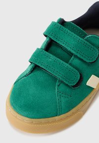 Sapato de camurça verde com duas correias de Velcro pretas, sola bege, bico arredondado e sutis detalhes de costura. Apresenta uma sola de borracha texturizada.