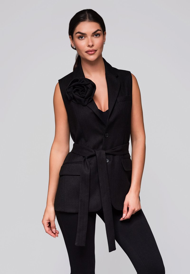 Zwarte mouwloze blazer met een grote stoffen roos op de linkerschouder, een ceintuur op de taille en twee voorzakken op een gladde, effen achtergrond.