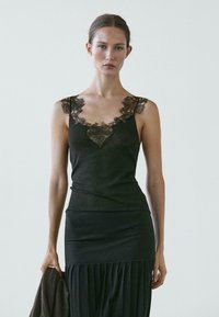 Donna che indossa una canottiera nera con rifiniture in pizzo e una gonna plissettata, tiene un capo scuro davanti a uno sfondo chiaro e uniforme.