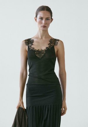 Donna che indossa una canottiera nera con rifiniture in pizzo e una gonna plissettata, tiene un capo scuro davanti a uno sfondo chiaro e uniforme.