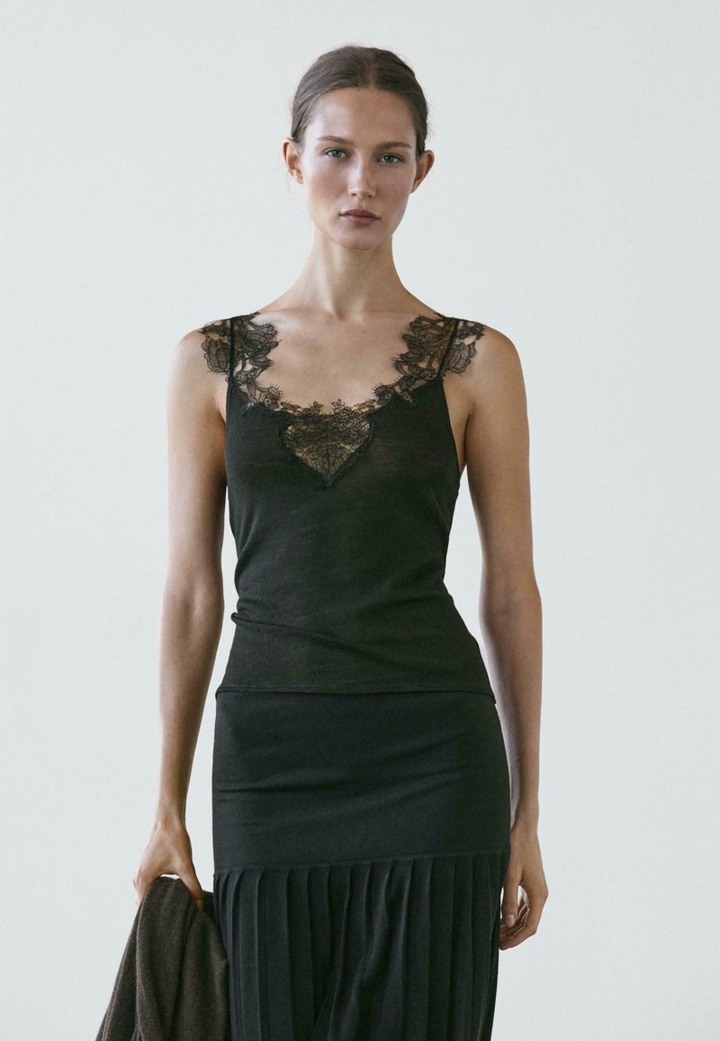 Donna che indossa una canottiera nera con rifiniture in pizzo e una gonna plissettata, tiene un capo scuro davanti a uno sfondo chiaro e uniforme.