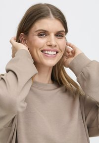 Sweatshirt beige clair à col rond et manches longues amples. Texture douce visible ; le modèle tient ses cheveux derrière les oreilles, souriant.