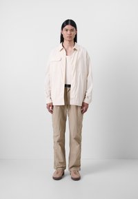 Beige, lazán illeszkedő ing, két elülső zsebbe, fehér ujjatlan póló fölött. Hozzá illő bézs nadrág texturált felülettel és világosbarna cipő.