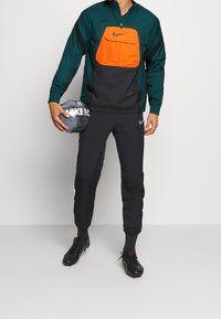 Hombre con chándal negro y verde de Nike sosteniendo un balón de fútbol negro de Nike, vestido con botas negras y de pie contra un fondo liso.
