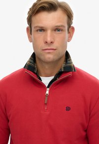 Rode ruime pullover met rits en een geruite kraag, voorzien van een klein geborduurd logo op de borst en een gladde, zachte textuur.