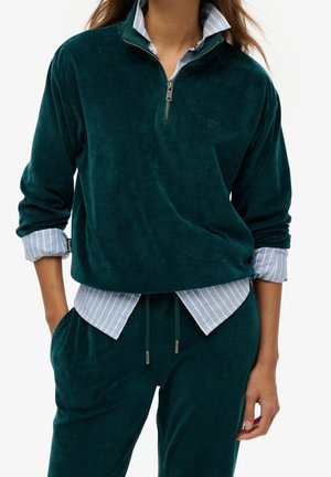 Persoon draagt een donker turquoise velours half-zip pullover en bijpassende broek over een lichtblauw gestreept overhemd met opgerolde mouwen.
