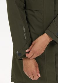 Olivgrön vattentät jacka med texturerad tyg, justerbara ärmslut och en logotyp som säger "W-PRO 10000" på ärmen.
