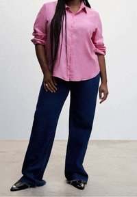 Personne avec de longues tresses portant une chemise rose à boutons, un jean bleu foncé à jambes larges, et des chaussures noires à bout pointu, debout sur un sol uni.