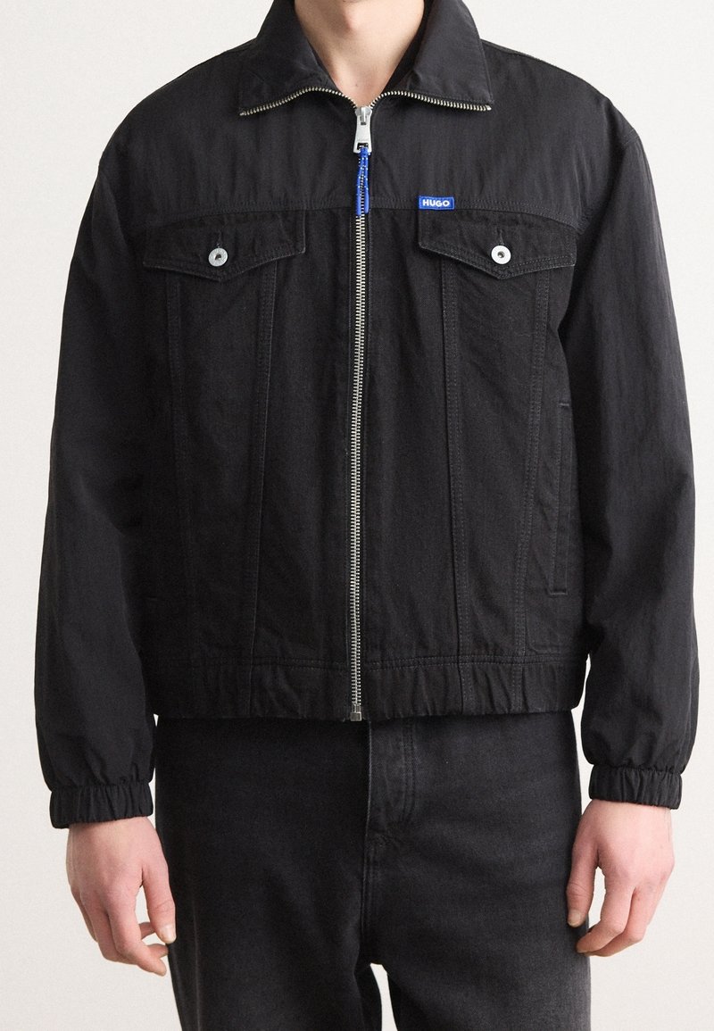 Giacca nera con zip, polsi elastici, tasche sul petto con pattina e bottoni, e un piccolo logo blu "HUGO" sul lato sinistro del petto.
