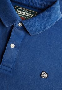 Polo bleu avec deux boutons blancs, un col côtelé, un logo "SD" brodé sur la poitrine, et une étiquette de marque "Superdry 1954" à l'intérieur du col.