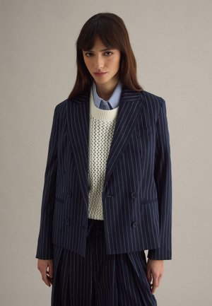 Frau trägt einen marineblauen, pinstripe doppelt geknöpften Blazer und eine Hose über einem weißen Strickpullover und einem blauen Hemd mit Kragen, steht vor einem schlichten Hintergrund.