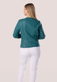 Giacca in pelle turchese con un design aderente, cuciture posteriori e maniche lunghe, abbinata a pantaloni bianchi, mostrata di schiena.