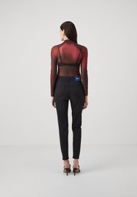 Rød og svart gjennomsiktig langermet topp paret med svarte tettsittende jeans. Har høy hals og merket bakpå. Modellen har på seg svarte hæler.