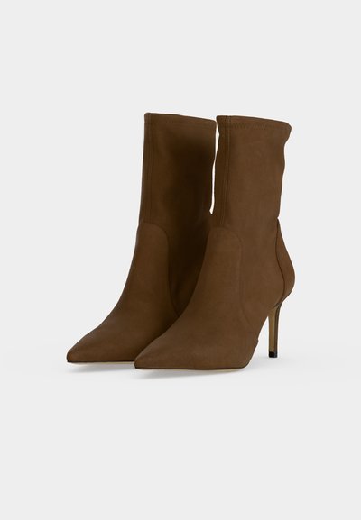 Bottines marron à talons hauts avec bout pointu et talons stilettos fins sur fond gris clair.