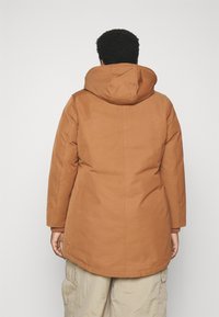 Abrigo parka marrón con capucha, que presenta una textura suave, corte recto y puños elásticos, llevado sobre pantalones de corte holgado de color claro.