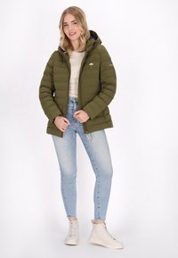 Olivgrön pufferjacka med huva, dragkedja och sidofickor, bärs över en beige topp och ljusblå jeans, tillsammans med beige sneakers.