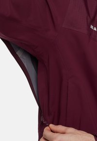 Giacca bordeaux realizzata in un tessuto leggero e morbido, con una tasca laterale con zip, un cordoncino regolabile e un pannello superiore testurizzato.