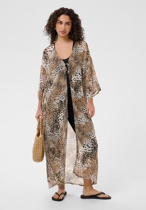 Nő átlátszó, állatmintás hosszú kimonót visel fekete ruházat felett, fonott táskát tart, és fekete szandált visel, egyszínű háttér előtt áll.