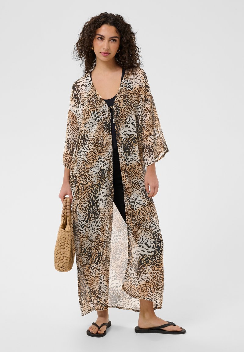 Femme portant un long kimono transparent à imprimé animalier sur une tenue noire, tenant un sac tissé et portant des sandales noires, debout sur un fond uni.