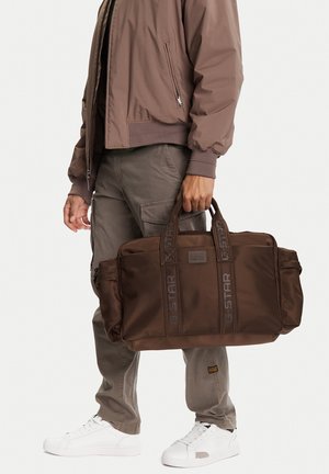 Persona con chaqueta marrón y pantalones cargo grises sosteniendo una bolsa de viaje G-Star grande de color marrón oscuro con asas dobles.