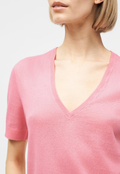 Femme portant un haut en maille rose à manches courtes et col en V, montrant le cou, la clavicule et une partie du visage sur un fond uni.