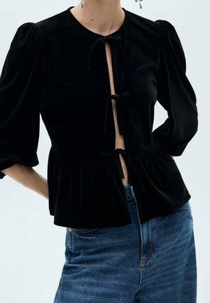Blouse en velours noir avec manches bouffantes et trois liens à nouer à l'avant, associée à un jean en denim bleu, visible du cou aux hanches.