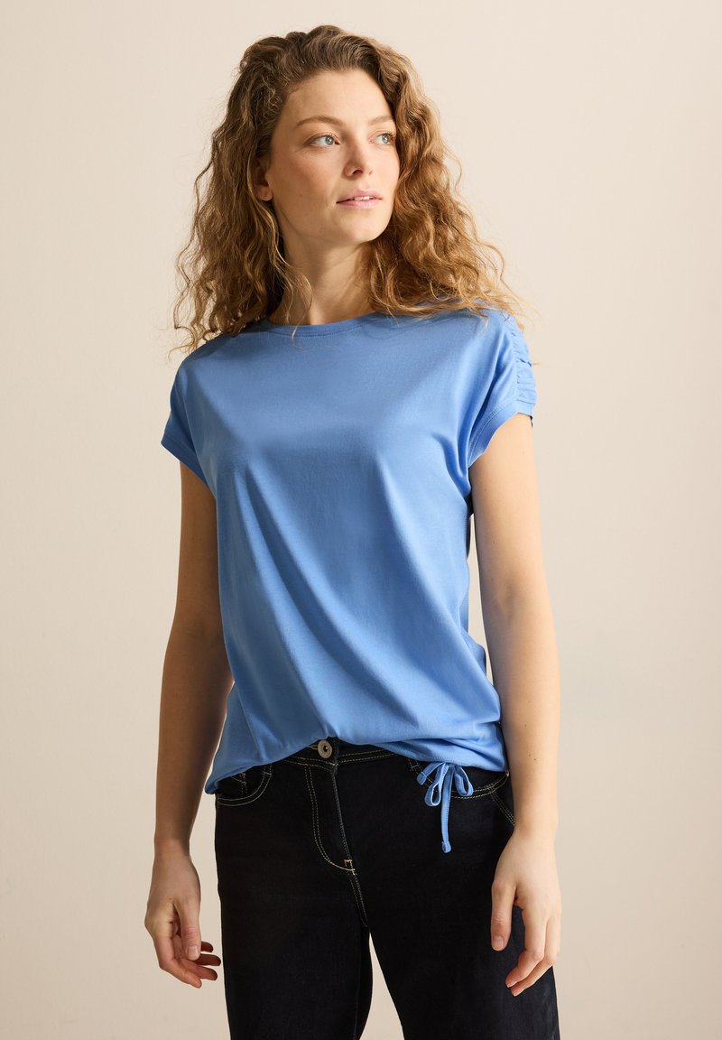 Camiseta de manga corta azul claro con escote redondo y detalle de hombro fruncido. Combinada con jeans oscuros que presentan un acento de cordón.