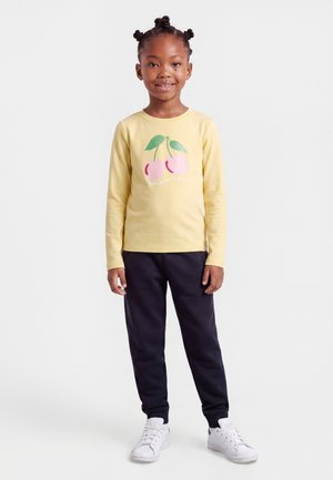 Fille aux cheveux tressés portant un t-shirt jaune à manches longues avec un motif cerise, pantalon noir et baskets blanches, debout et souriante.