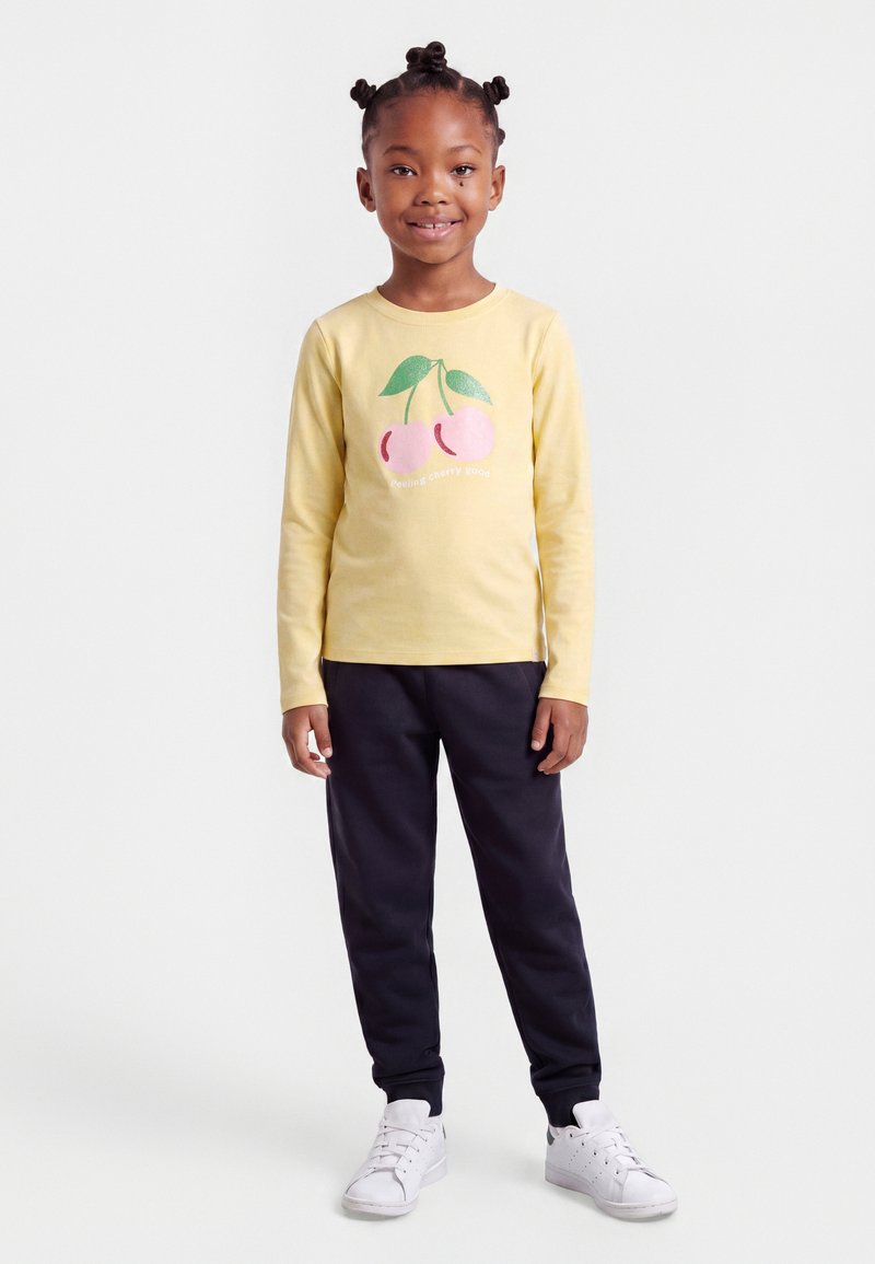 Fille aux cheveux tressés portant un t-shirt jaune à manches longues avec un motif cerise, pantalon noir et baskets blanches, debout et souriante.