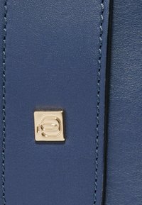 Bolso de mano de cuero azul con un logo cuadrado dorado, textura suave, costuras detalladas y un diseño elegante.