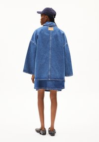 Oversized denim jacket with wide sleeves, blue color, featuring a back label patch and layered hem. Worn with black slip-on shoes.