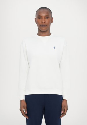 Mujer con cabello corto, vistiendo un sudadera blanca de manga larga y pantalones azul oscuro, de pie contra un fondo claro y liso.