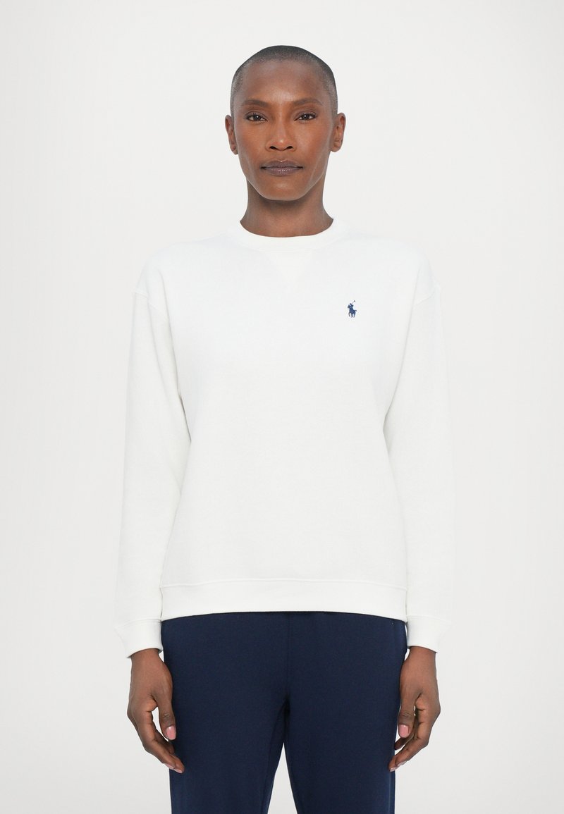 Femme aux cheveux courts portant un sweatshirt blanc à manches longues et un pantalon bleu foncé, se tenant devant un fond clair uniforme.