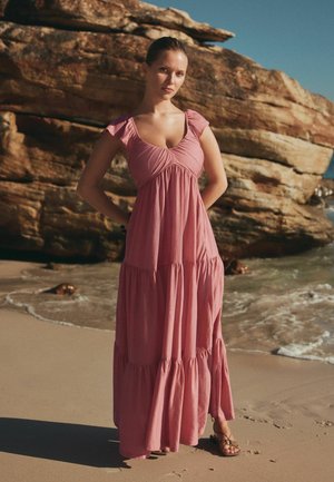 Woman in a pink tiered maxi dress and sandals stands on sandy beach with rocky cliffs and ocean waves in the background.