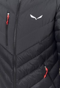 Gros plan sur une veste noire matelassée avec une fermeture éclair centrale et des cordons rouges aux poches poitrine et latérales, arborant un logo angulaire blanc.