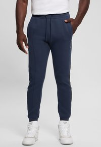 Pantaloni jogger tapered blu navy in un morbido tessuto, con vita regolabile tramite cordoncino, strisce laterali e tasche inclinate, abbinati a sneaker alte bianche.