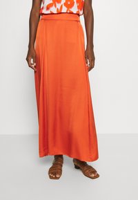 Maxirock aus leuchtend orangefarbenem Satin mit glatter Textur, ausgestattet mit hoher Taille und fließendem Design, kombiniert mit braunen Riemchensandalen.