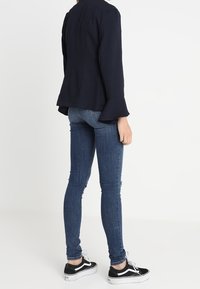 Námořnická bunda s rozšířenými rukávy, spárovaná s úzkými tmavě modrými džíny a černými teniskami s bílou podrážkou. Minimalistický design, hladká textura.