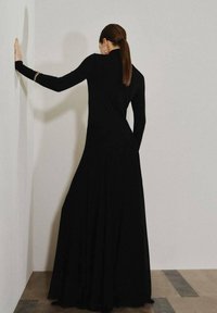 Largo vestido negro con mangas largas, ajustado en la parte superior y fluido en la parte inferior. Textura suave, diseño minimalista, adornado con detalles en oro.