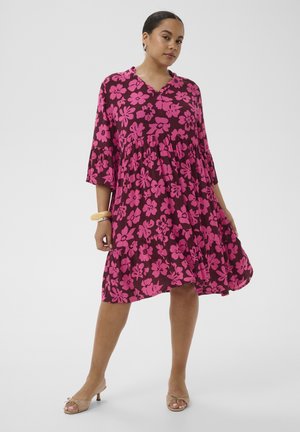 Kobieta ubrana w luźną, sięgającą kolan fioletową sukienkę w różowy kwiatowy wzór, z rękawami do łokcia, w beżowe sandały na obcasie i z bransoletkami na jednym nadgarstku.